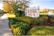 Outdoor view of a landscaped area with green bushes and trees surrounding a white sign that reads 'Autumn House' in red letters, with a sidewalk on the left side and a clear sky above.