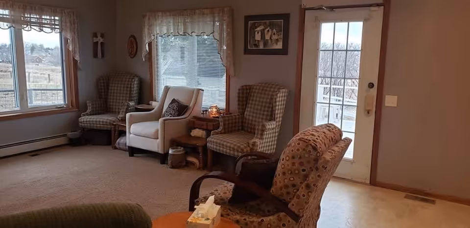 A cozy living room area with several upholstered armchairs arranged near windows with lace curtains and a glass door. The room has beige walls, carpeted flooring, and a small wooden side table with decorative items. Outside the windows, a rural landscape is visible.