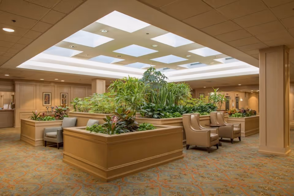 Indoor seating area with multiple beige armchairs arranged around large wooden planters filled with various green plants. The ceiling features a large recessed light panel with square cutouts, illuminating the space. The walls are paneled in light wood, and the carpet has a patterned design in muted tones.