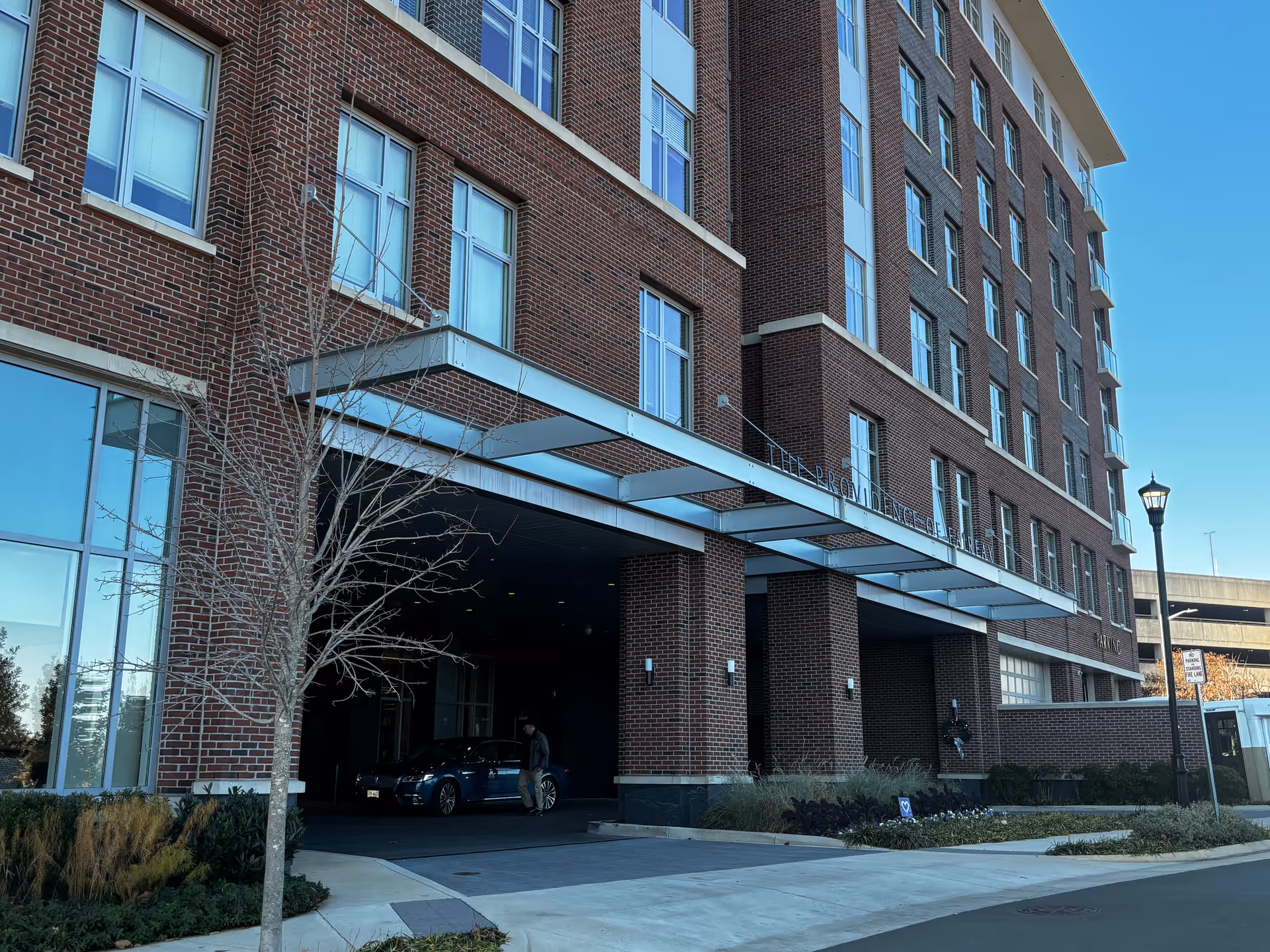 Exterior view of a multi-story brick building with large windows and a covered driveway entrance. A person is walking near a parked car under the entrance canopy. The building has a sign that reads 'The Providence Fairfax'. There is a street lamp and some landscaping including a small tree and bushes near the sidewalk.