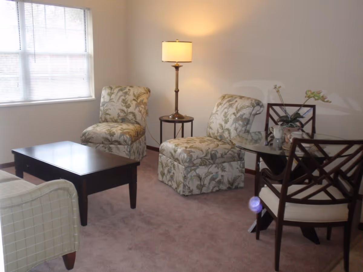 A cozy living room area with two floral-patterned armchairs, a small round side table with a lamp, a dark wooden coffee table, and a glass-top dining table with two wooden chairs. A window with blinds allows natural light into the room.