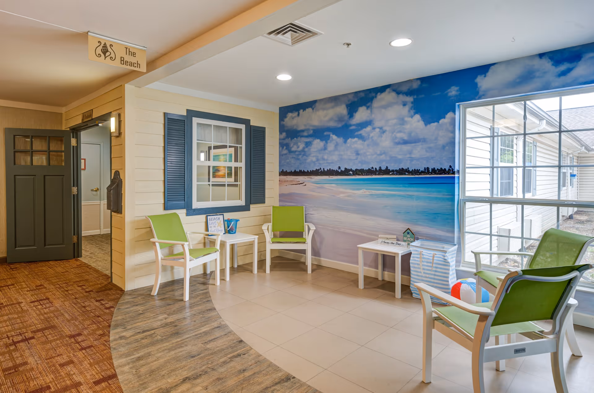 A bright indoor seating area with green chairs and small white tables. The wall features a large mural of a beach scene with blue skies, white clouds, and turquoise water. There is a window showing the exterior of the building. A sign above the doorway reads 'The Beach.'