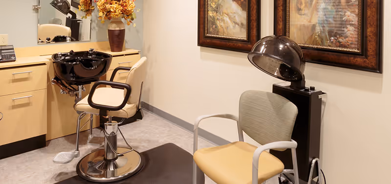 Interior view of a hair salon area in a memory care facility featuring a salon chair in front of a black hair washing sink, a beige chair with a hair dryer hood, a countertop with a telephone and a vase with flowers, and framed artwork on the wall.