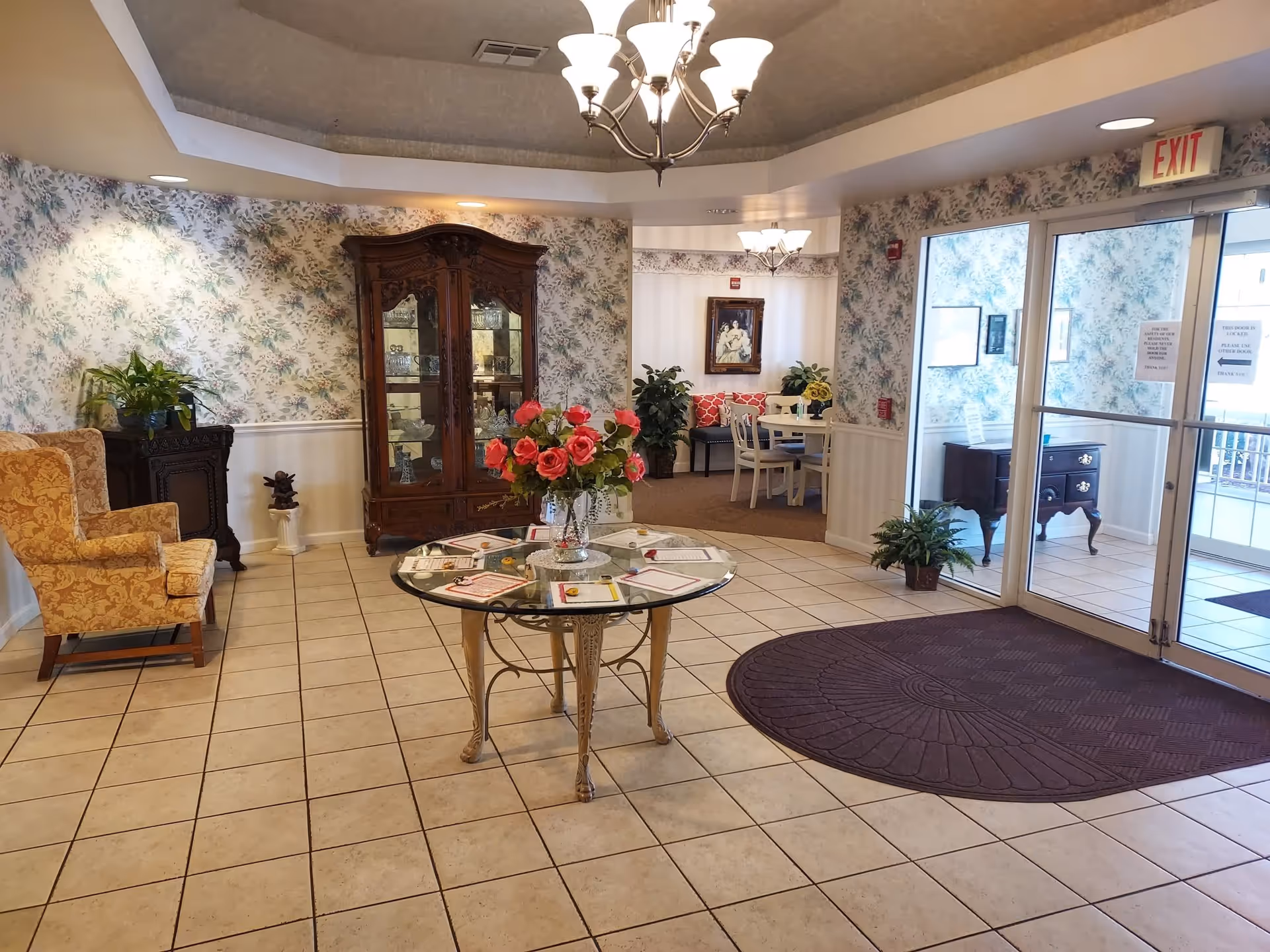 An entrance lobby area with tiled flooring and floral wallpaper. A round glass table with a vase of pink flowers is centered on the floor. To the left, there is a yellow patterned armchair and a dark wooden cabinet with a plant on top. In the background, a wooden china cabinet displays glassware. To the right, glass double doors lead outside, with a dark floor mat in front of them. A small seating area with a round table, chairs, and red cushions is visible through an open doorway. Ceiling lights and an exit sign are also visible.