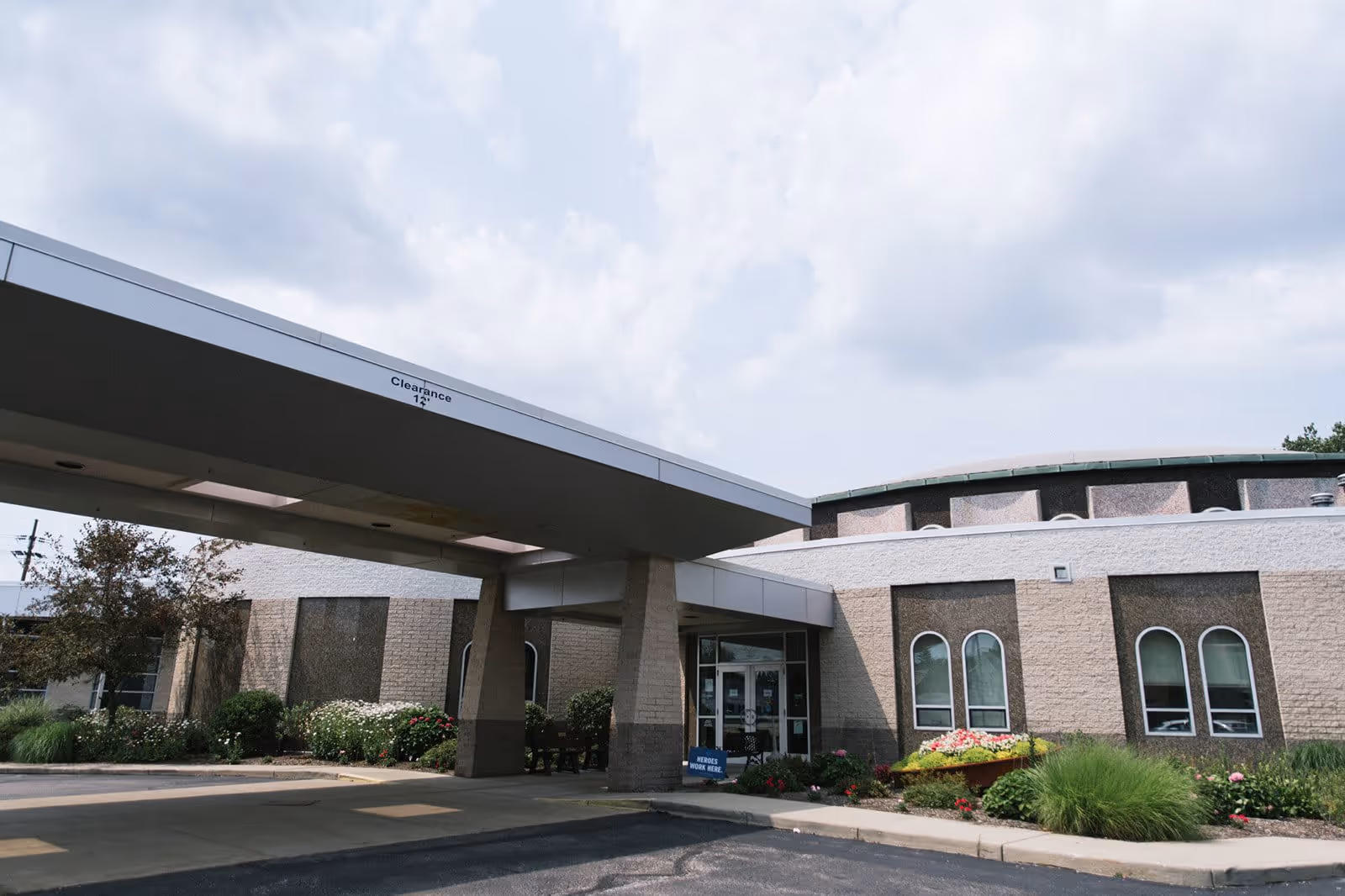 Exterior view of Light of Hearts Villa showing the entrance with a covered drop-off area, landscaped bushes and flowers, and a partly cloudy sky.
