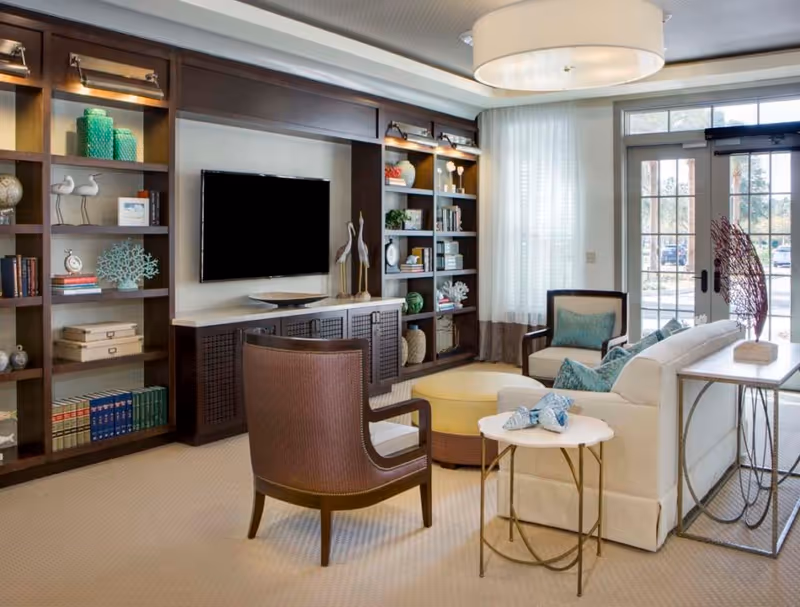 A bright communal living room with built-in bookshelves, a wall-mounted TV, sofa and armchairs, and large windows letting in natural light.