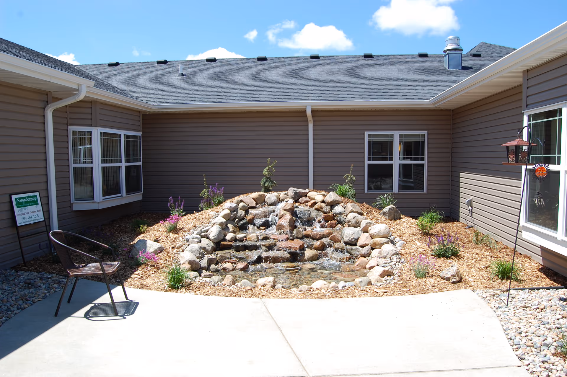 Outdoor courtyard area with a small rock waterfall feature surrounded by mulch and plants, enclosed by a building with beige siding and several windows. There is a single metal chair on the concrete patio and a bird feeder hanging near a window.