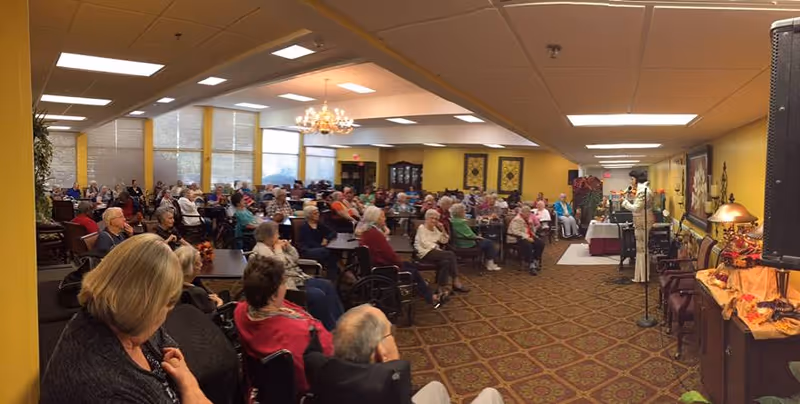 A large group of elderly people seated at tables and chairs in a spacious room with yellow walls and patterned carpet, attentively listening to a woman speaking at a microphone near the front. The room is well-lit with ceiling lights and has windows along one side. There are decorative elements including a chandelier, framed artwork, and a table with autumn-themed decorations.