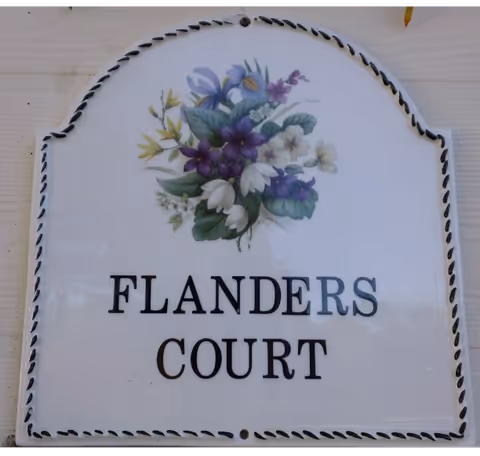 Decorative white ceramic plaque with a floral motif that reads "FLANDERS COURT" mounted on siding.