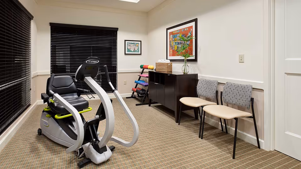 A small exercise room with a recumbent exercise bike, a rack of colorful dumbbells, two chairs with patterned upholstery, a dark wooden cabinet with a vase and tissue box on top, and framed artwork on the walls. The room has beige walls, carpeted floor, and windows with black blinds.