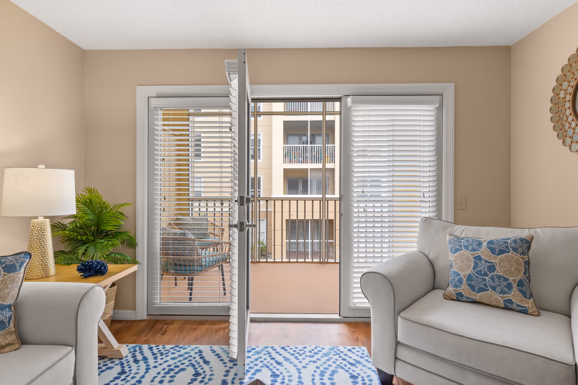 A cozy living room with beige walls and wooden flooring. There are two light gray armchairs with blue and beige patterned cushions. A wooden side table with a textured lamp and a green plant is placed next to one armchair. White blinds cover the glass sliding door that opens to a small balcony with outdoor chairs. The balcony overlooks another building with balconies.