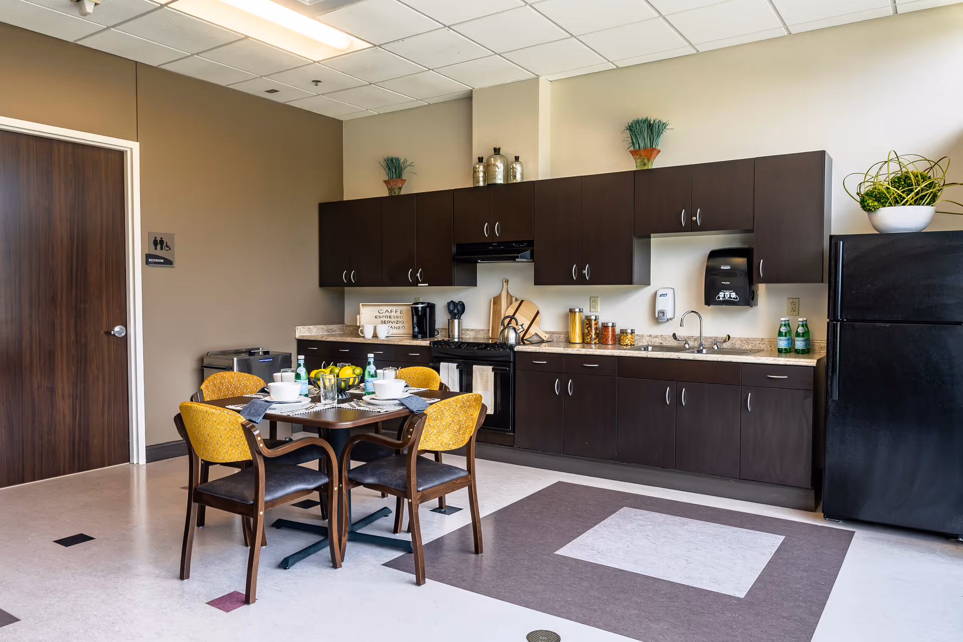 A modern kitchen area with dark wood cabinets, a black refrigerator, and a black stove. A small dining table with four yellow cushioned chairs is set with white bowls, plates, and cups. The countertop has various kitchen items including a coffee maker, jars, and decorative plants. A door labeled as a restroom is visible on the left side of the image.