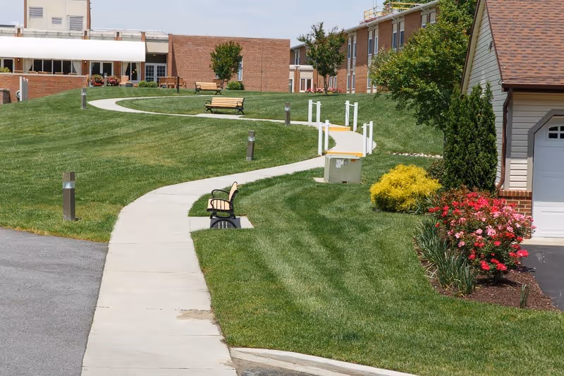 A winding concrete pathway through a well-maintained grassy area with benches along the path. There are bushes with red and yellow flowers near a building with a garage door. The background shows brick buildings and small trees under a clear sky.