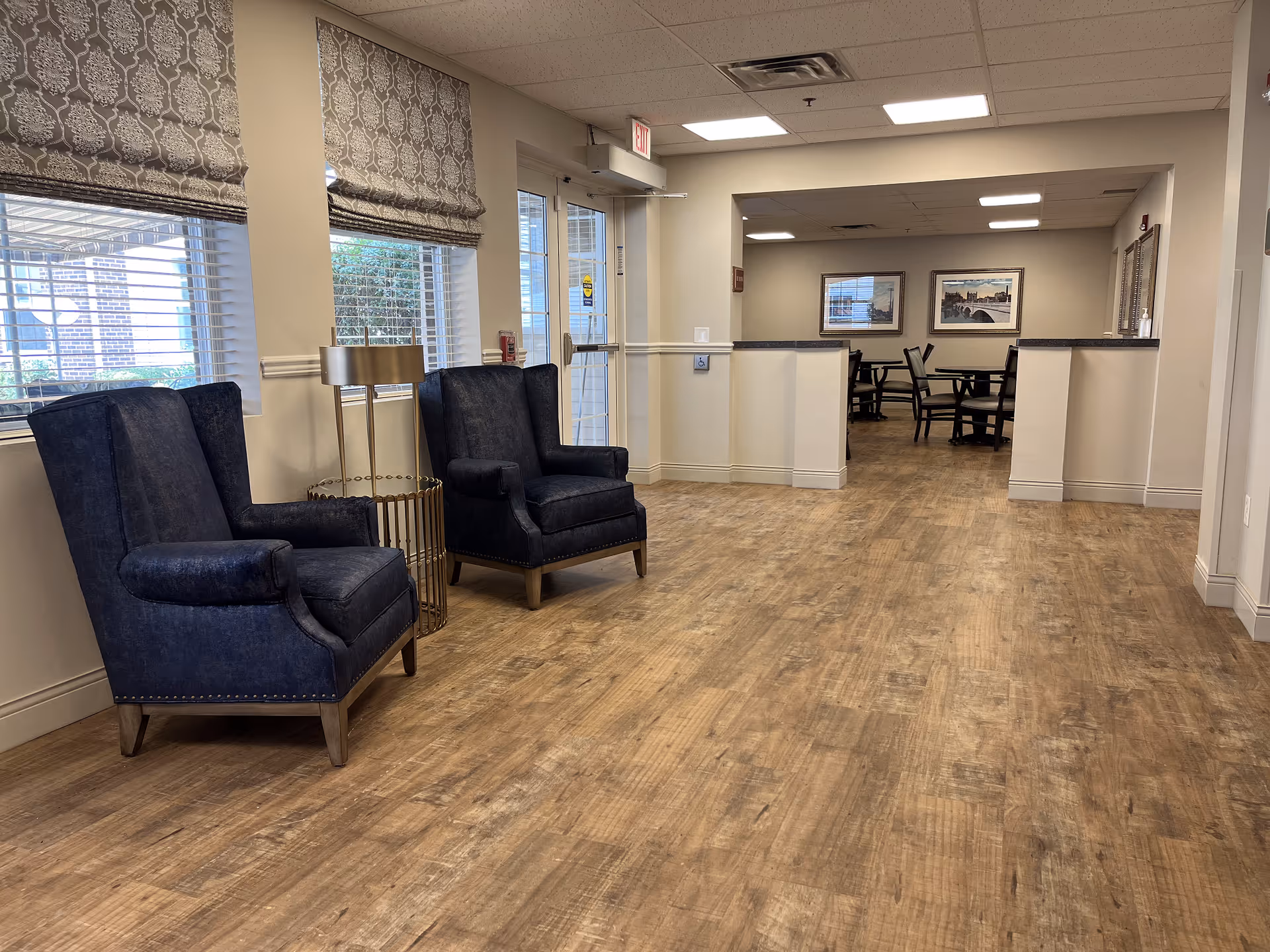 Interior view of a senior living facility showing a hallway with two dark blue upholstered armchairs on the left near windows with patterned Roman shades. The floor is wood-style, and the hallway leads to a dining area with tables and chairs. The walls are light-colored, and the ceiling has recessed lighting.