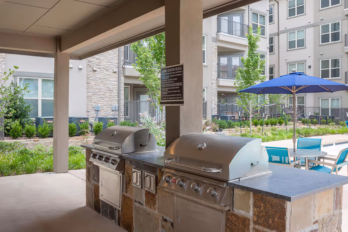 Outdoor covered grilling area with two stainless steel gas grills built into a stone countertop. In the background, there are apartment buildings, green landscaping, a blue patio umbrella, and outdoor seating with tables and chairs.