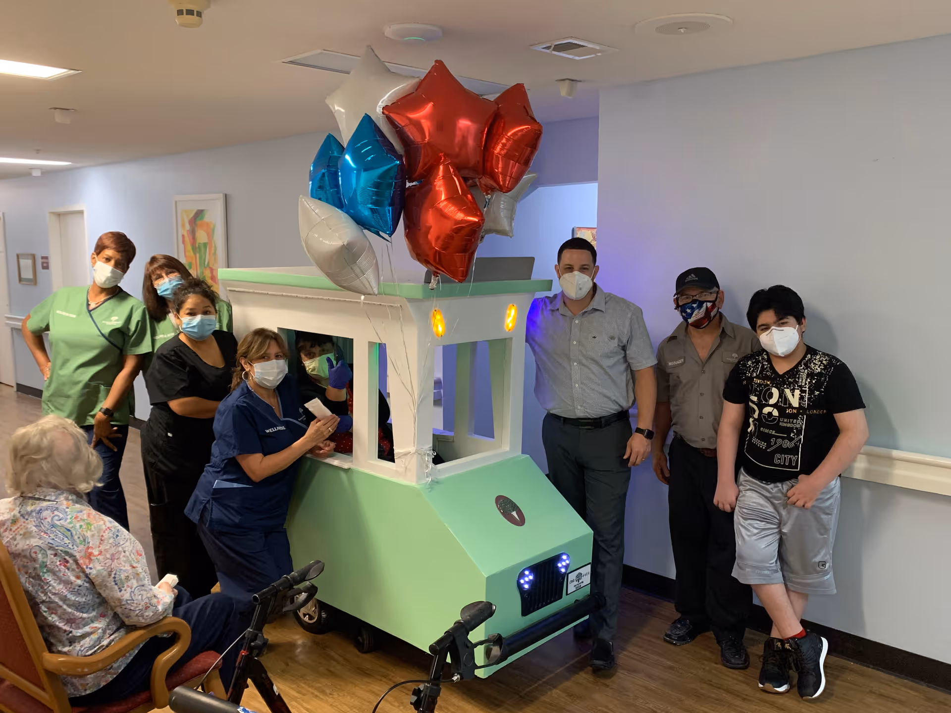A group of seven people wearing face masks stand around a small green and white cart decorated with red, white, and blue star-shaped balloons in a hallway. An elderly woman with white hair sits in a chair facing the group. The setting appears to be inside a senior living facility.