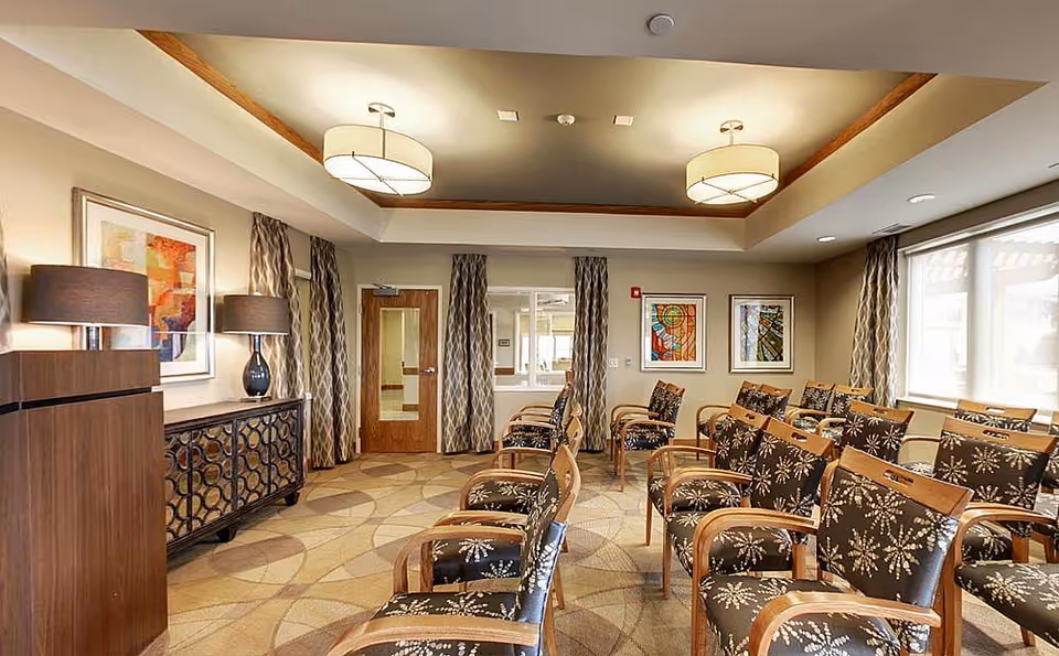 A well-lit meeting or activity room with multiple rows of wooden chairs featuring floral patterned cushions. The room has large windows with patterned curtains, two ceiling light fixtures, framed abstract artwork on the walls, and a wooden cabinet with two table lamps on the left side.