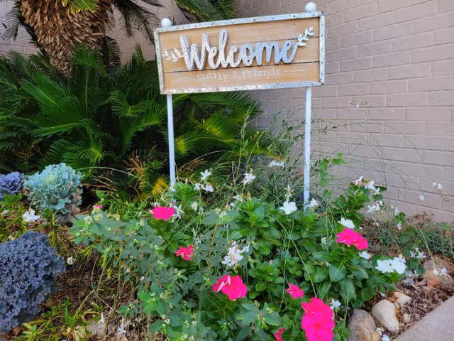 A garden area with various green plants and colorful flowers including pink and white blooms. Behind the plants is a wooden sign on white metal posts that reads 'Welcome family & friends'. The background includes a brick wall and some palm leaves.