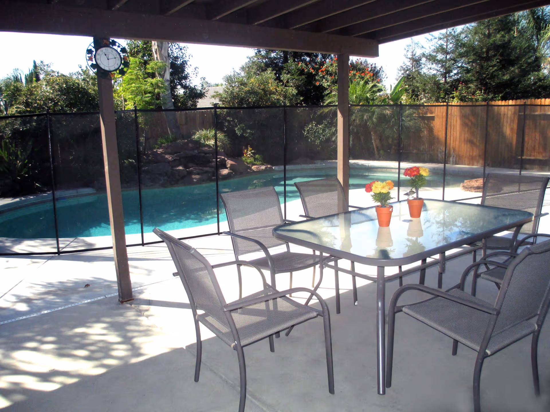 Outdoor patio area with a glass table and six metal mesh chairs under a covered structure. Three small potted plants with colorful flowers are placed on the table. In the background, there is a fenced swimming pool surrounded by trees and a wooden fence.