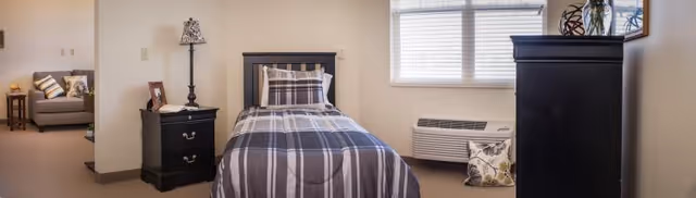 Small furnished bedroom with a single bed in plaid bedding, a nightstand and lamp, a dresser, a window with blinds, and a view into an adjacent sitting area.