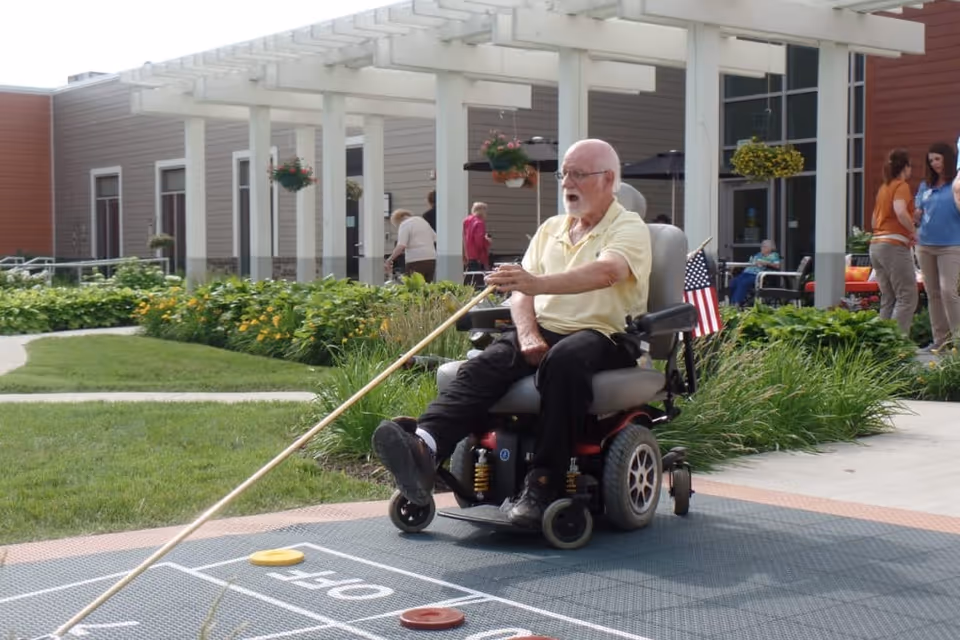 An elderly man in a motorized wheelchair playing shuffleboard outdoors at a senior living facility. He is holding a long shuffleboard cue and aiming at the shuffleboard court. The background shows a garden with green plants and flowers, a white pergola, and several other people walking and sitting near the building.