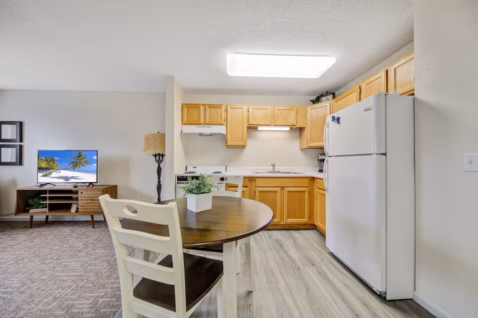 Interior view of a senior living facility apartment showing a small kitchen with wooden cabinets, a white refrigerator, a stove, and a sink. In the foreground, there is a round wooden dining table with two white chairs and a small potted plant on the table. To the left, a living area is partially visible with a TV on a wooden stand displaying a beach scene and a floor lamp beside it.