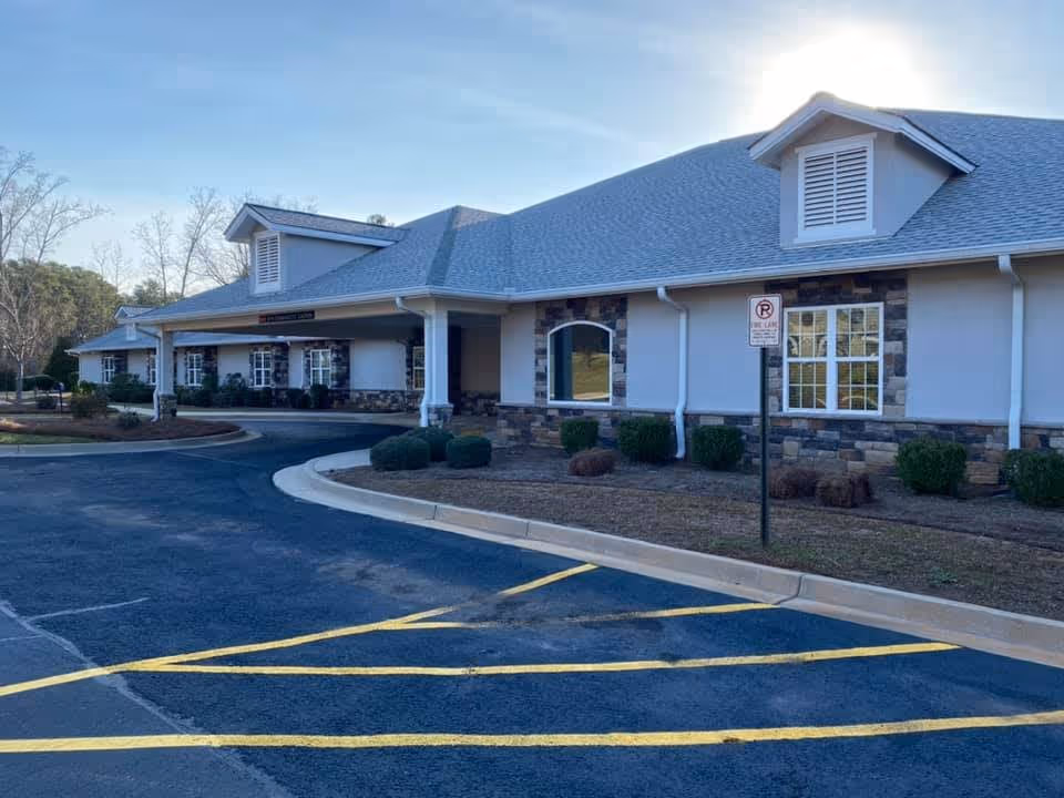 Single-story senior living/memory care building with a covered drop-off entrance, paved parking area with yellow lines, and low landscaping.