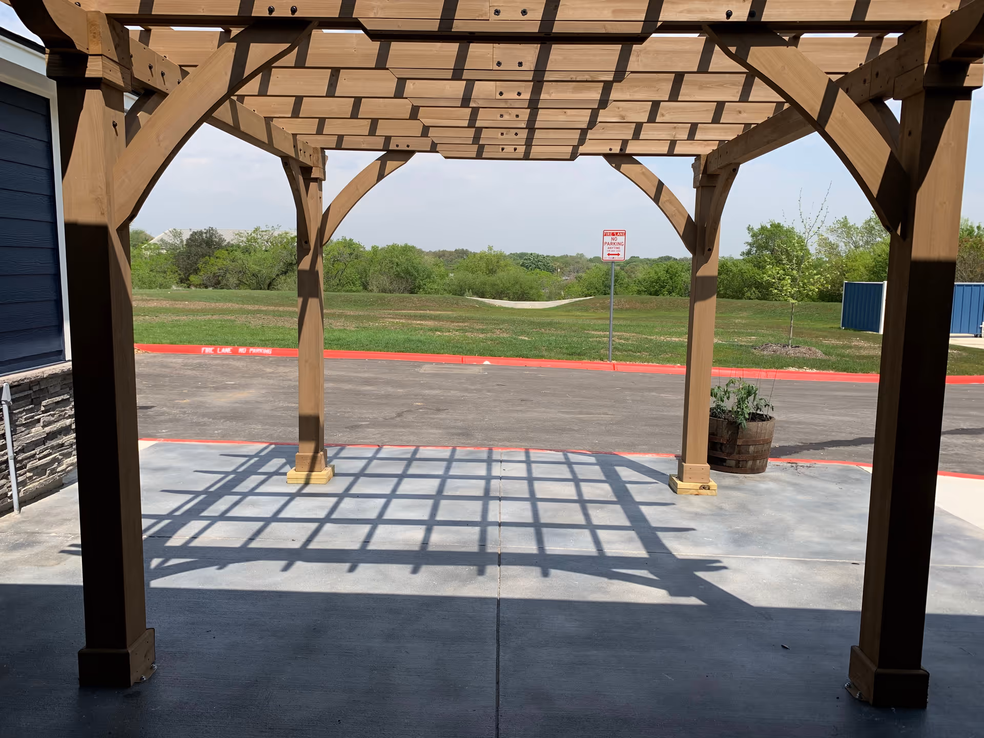 View from under a wooden pergola structure looking out onto a paved area with a red curb and a grassy field beyond. There is a small barrel planter with plants on the right side and a 'No Parking' sign in the distance.