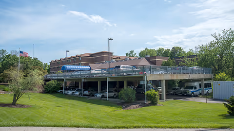 Exterior view of Western Hills Retirement Village showing a brick multi-story building with an elevated parking deck, cars and a bus beside a manicured lawn.