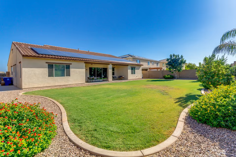 Well-maintained backyard with a green lawn, curved stone borders, flower beds, and a single-story house with solar panels and a covered patio.