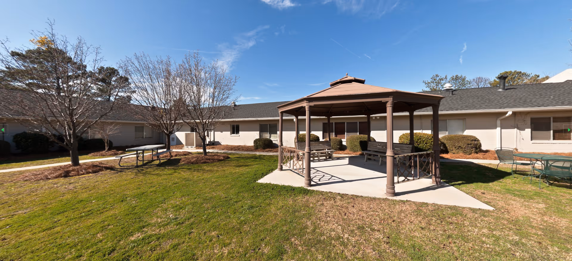 Outdoor courtyard area at a senior living facility with a brown gazebo featuring benches inside, surrounded by grass, leafless trees, and picnic tables. The building with beige walls and multiple windows forms a U-shape around the courtyard under a clear blue sky.