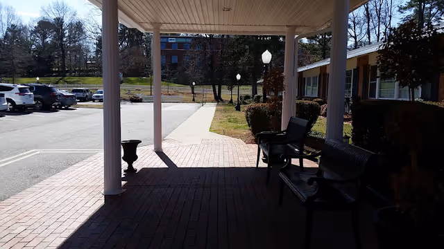 Covered porte-cochère with benches, a brick walkway, lamp posts, and a parking lot in front of a low brick building.