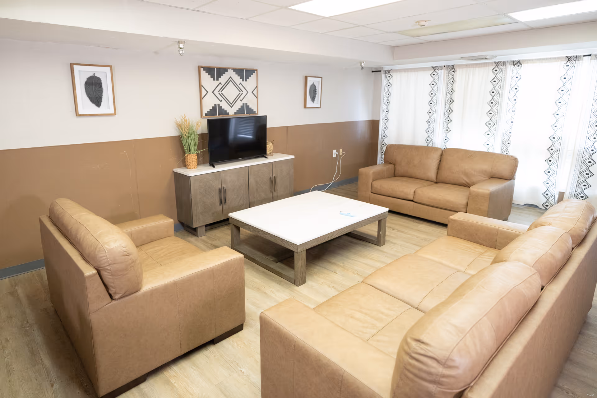 A living room with three tan leather sofas arranged around a rectangular coffee table. A TV sits on a cabinet against the wall, flanked by two framed pictures and a plant. Light-colored curtains with a geometric pattern cover the windows, and the walls are painted half white and half brown.