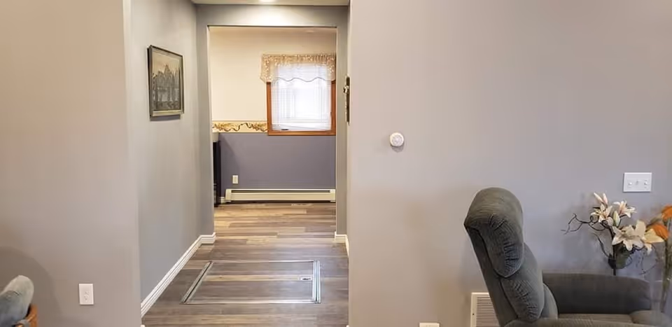 Interior view of a senior living facility showing a hallway with wood flooring leading to a room with a window covered by a sheer curtain. On the right side, there is a comfortable recliner chair next to a small table with a vase of flowers. The walls are painted light gray and decorated with a framed picture.