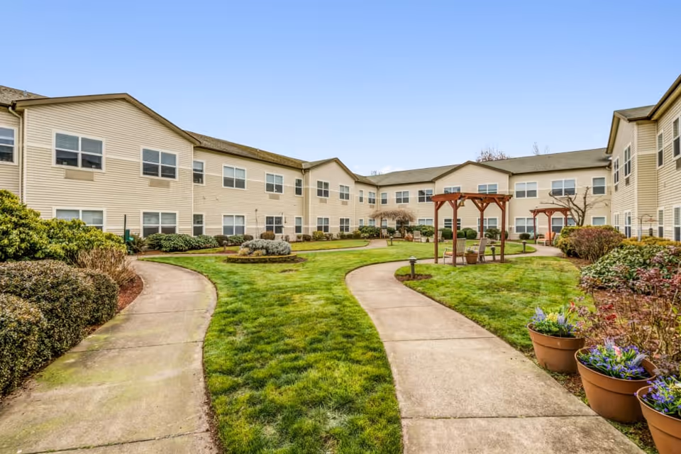 Courtyard of a two-story beige senior living building with curved concrete pathways, grassy lawn, pergolas, and potted plants.