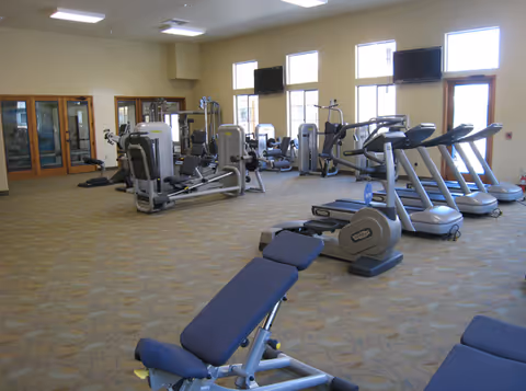 A spacious fitness room with various exercise equipment including treadmills, stationary bikes, weight machines, and benches. The room has large windows allowing natural light to enter and multiple wall-mounted televisions.