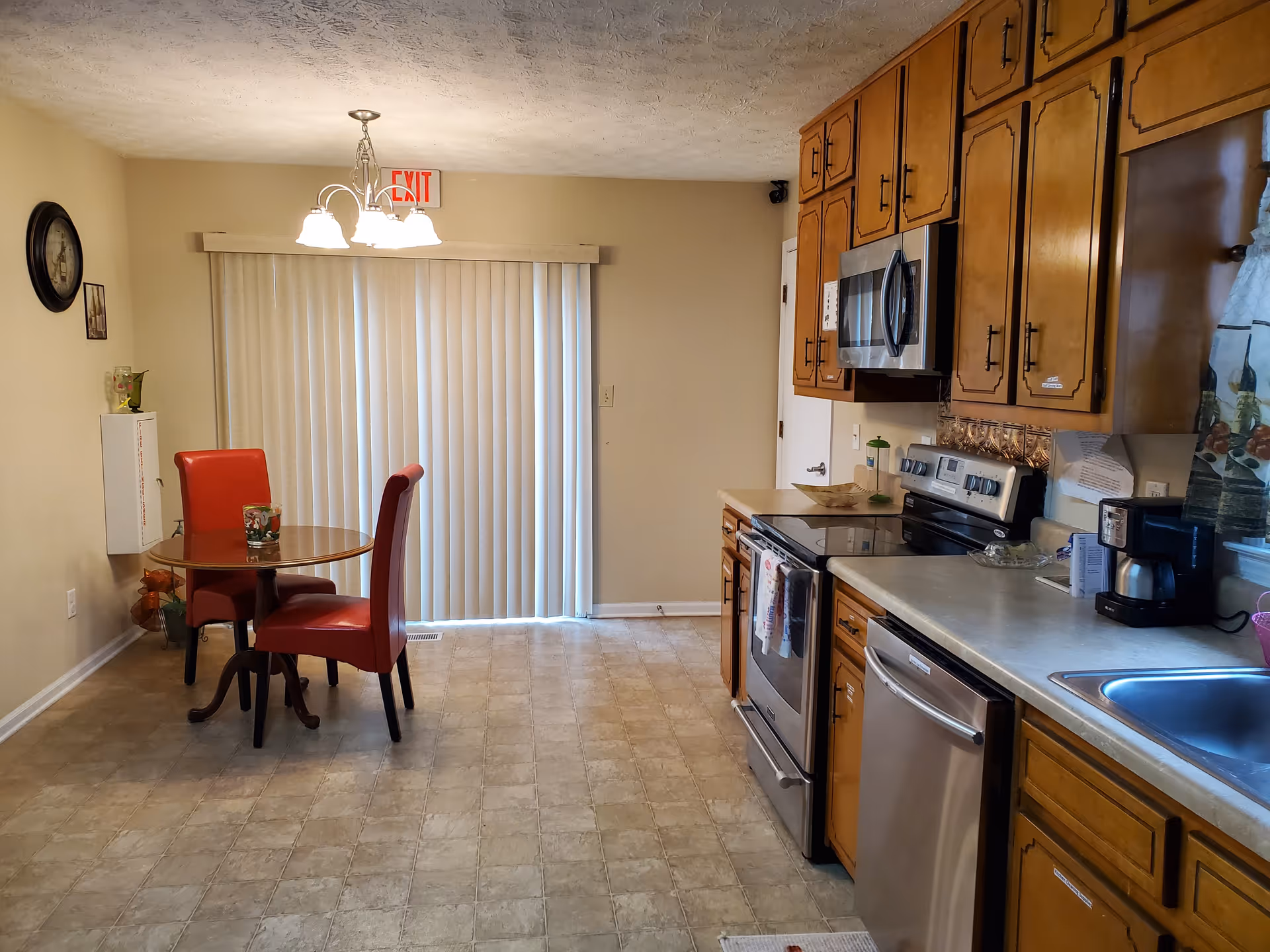 A residential kitchen with wooden cabinets, stainless steel appliances, a sink and a small round dining table with two red chairs in front of vertical blinds.