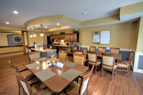 Large communal dining room with multiple wooden tables and chairs and an open kitchen area under warm lighting.