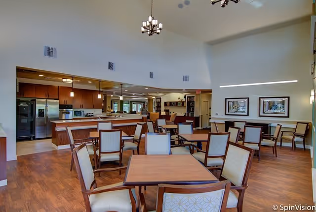 Spacious communal dining area with multiple wooden tables and chairs and a kitchen counter under high ceilings.