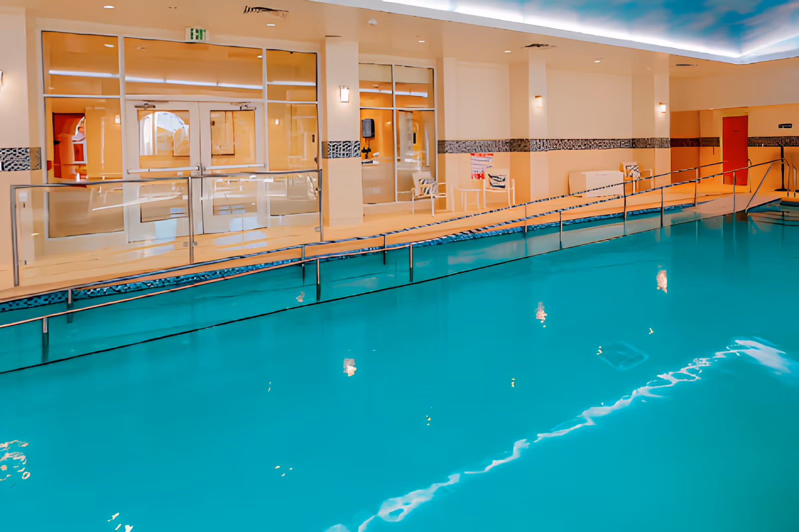 Indoor swimming pool with clear blue water, a ramp with handrails for accessibility, beige walls with decorative tile trim, glass doors and windows along one side, and ceiling lights reflecting on the water surface.