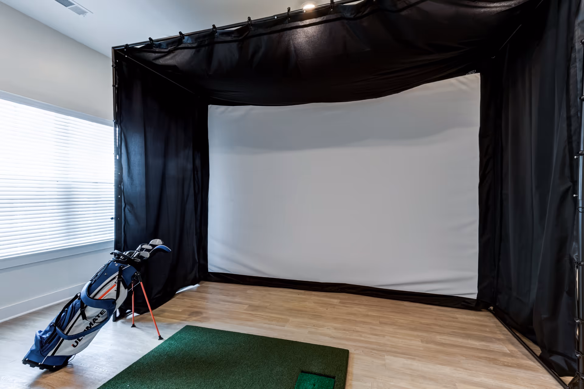 Indoor golf simulator setup with a large white screen surrounded by black curtains, a green artificial turf mat on the wooden floor, and a golf bag with clubs standing nearby next to a window with blinds.