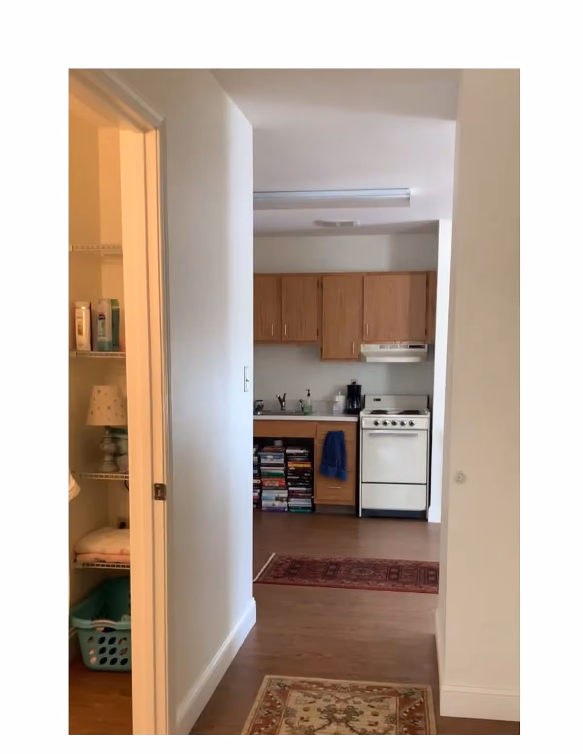 View down a hallway into a small kitchen area with wooden cabinets, a white stove, and a countertop with a coffee maker and soap dispenser. To the left, there is an open door revealing shelves with toiletries, a lamp, folded towels, and a laundry basket. The floor is wooden with two rugs placed along the hallway and kitchen entrance.