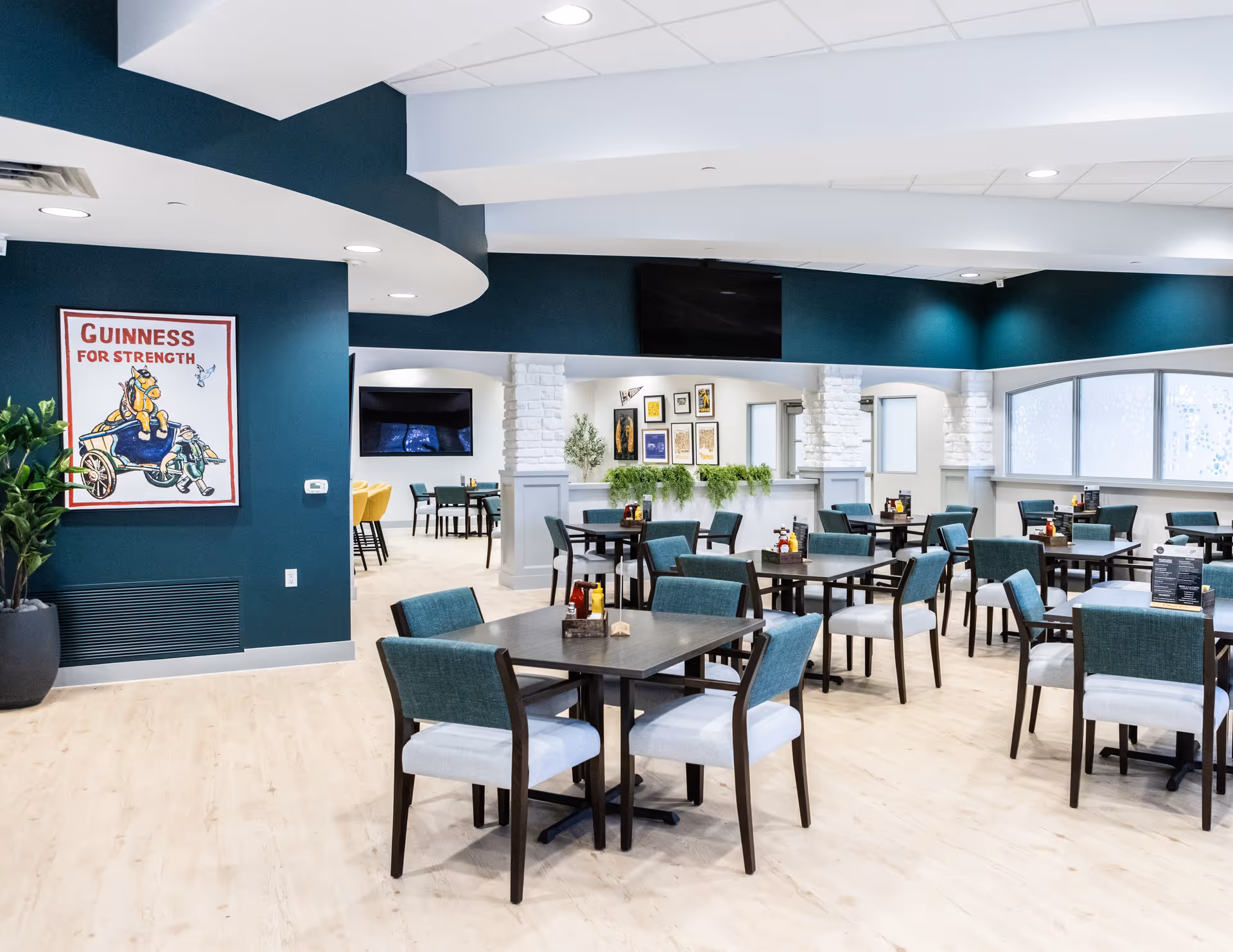 A bright and modern dining room with multiple square tables and teal cushioned chairs. The walls are painted teal and white with decorative white brick columns. There is a Guinness for Strength poster on one wall, several framed pictures on another, and a large flat-screen TV mounted above an archway. The floor is light wood, and there are condiment holders on each table.