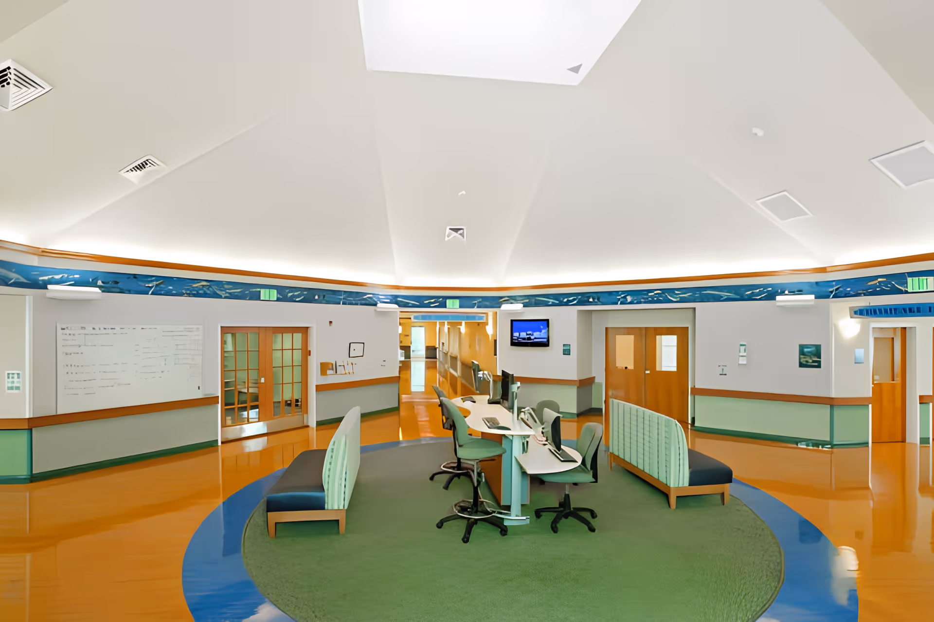 A spacious, well-lit interior area with a circular green carpet in the center, featuring a workstation with multiple chairs and computer monitors. Surrounding the workstation are cushioned benches with green upholstery. The walls are light-colored with wooden trim and a decorative blue border near the ceiling. Several doors and a hallway are visible in the background.