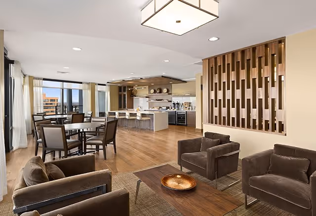 A spacious and modern senior living common area featuring a seating area with four brown armchairs around a wooden coffee table with a decorative bowl. In the background, there is a dining area with tables and chairs near large windows letting in natural light. The kitchen area includes a long island with bar stools and open shelving with kitchenware. The room has wooden flooring and a decorative wooden partition wall.