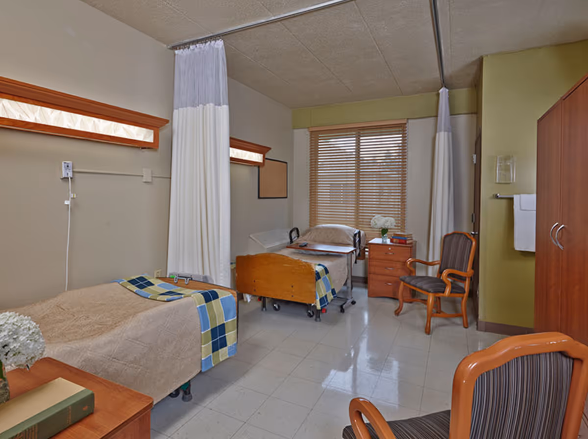 A shared bedroom in a healthcare facility with two hospital beds separated by a white privacy curtain. Each bed has a beige blanket and a plaid throw. There is a wooden nightstand with books and a flower vase between the beds. The room has a window with wooden blinds, two wooden chairs with striped cushions, a wooden wardrobe, and a towel rack with a white towel.