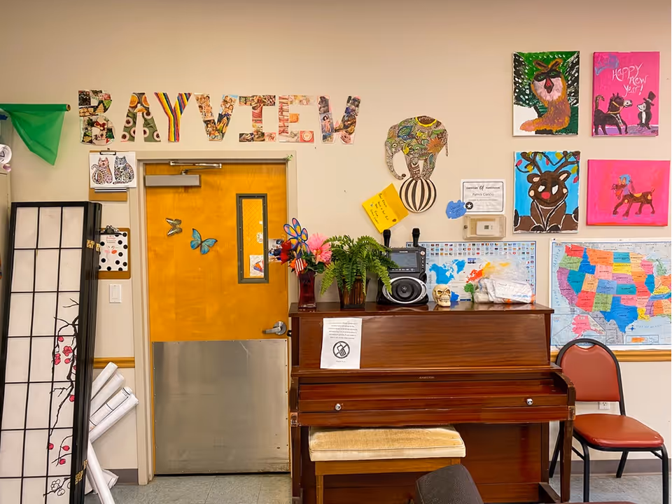 Interior room with a wooden piano against the wall, decorated with a vase of flowers, a fern plant, a skull, and a radio. Above the piano, colorful artwork and maps are displayed on the wall. The word 'BAYVIEW' is spelled out in large, decorated letters above a wooden door with butterfly decorations. A red chair is placed next to the piano, and a folding screen with a floral design is on the left side.