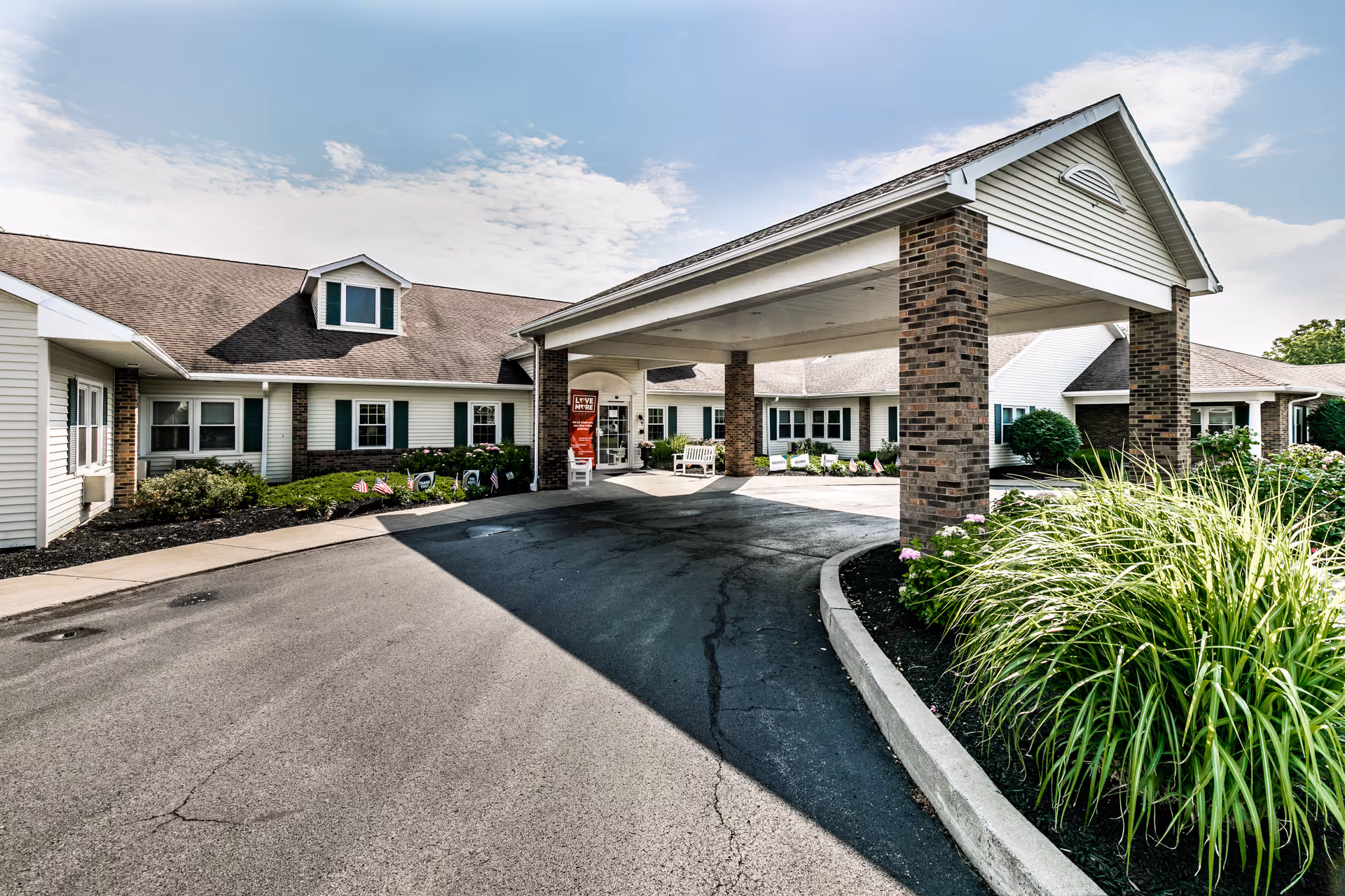 Front entrance of Keepsake Village at Greenpoint showing a covered porte-cochere with brick columns, driveway, and landscaped beds.