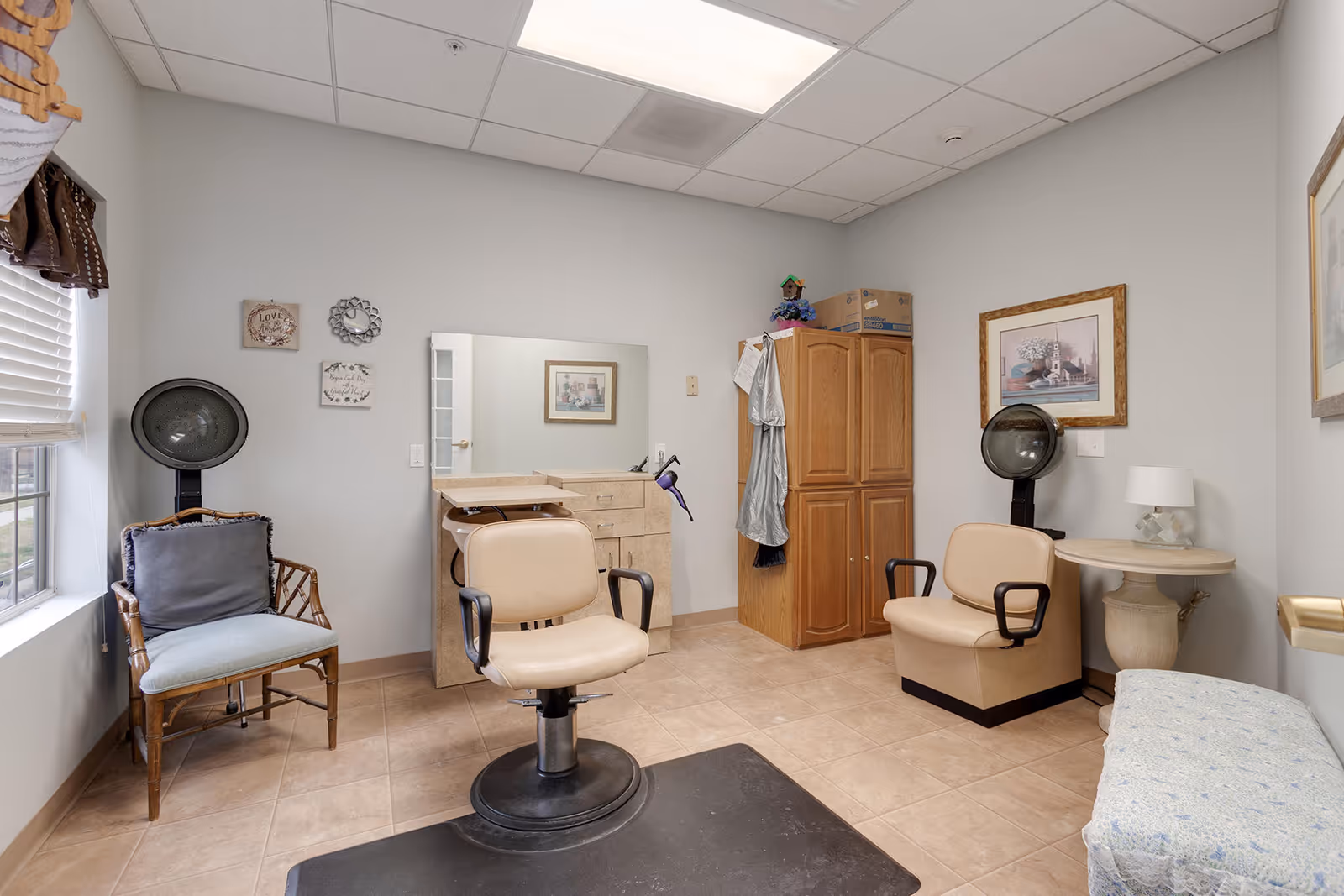 A small hair salon room with two beige salon chairs, two hair dryers, a wooden cabinet, a mirror above a counter with drawers, a wicker chair with a cushion near a window with blinds, framed artwork on the walls, and a small round table with a lamp.