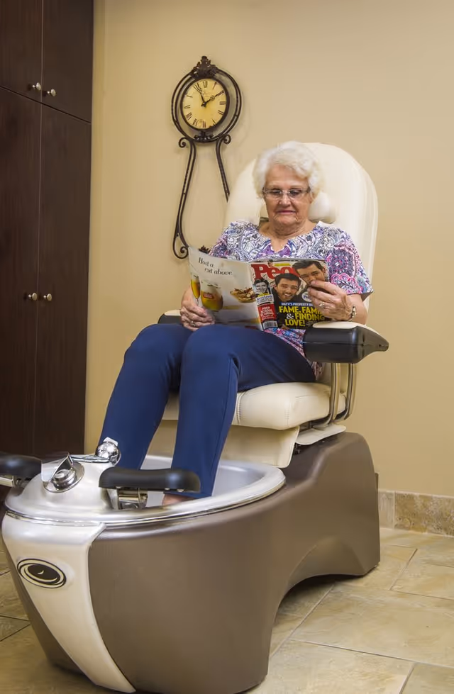 An elderly woman with white hair and glasses is sitting in a pedicure spa chair, reading a magazine. The chair has a foot bath basin at the bottom. Behind her is a wall clock with Roman numerals and a wooden cabinet to the left.